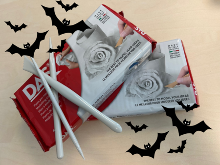 Två paket lufttorkande lera, tre modelleringsverktyg och tecknade svarta fladdermöss på ett ljust bord.