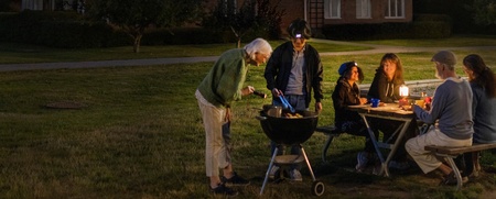 En grupp människor grillar och sitter vid ett bord upplyst av batterilampa utomhus på kvällen framför ett flerbostadshus.