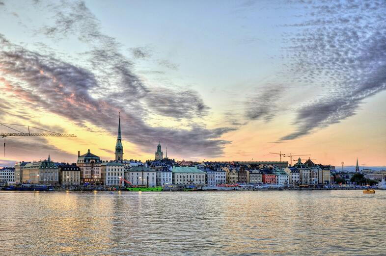Stadssilhuett vid vattnet i solnedgång, med färgglada byggnader och dramatiska moln på himlen.